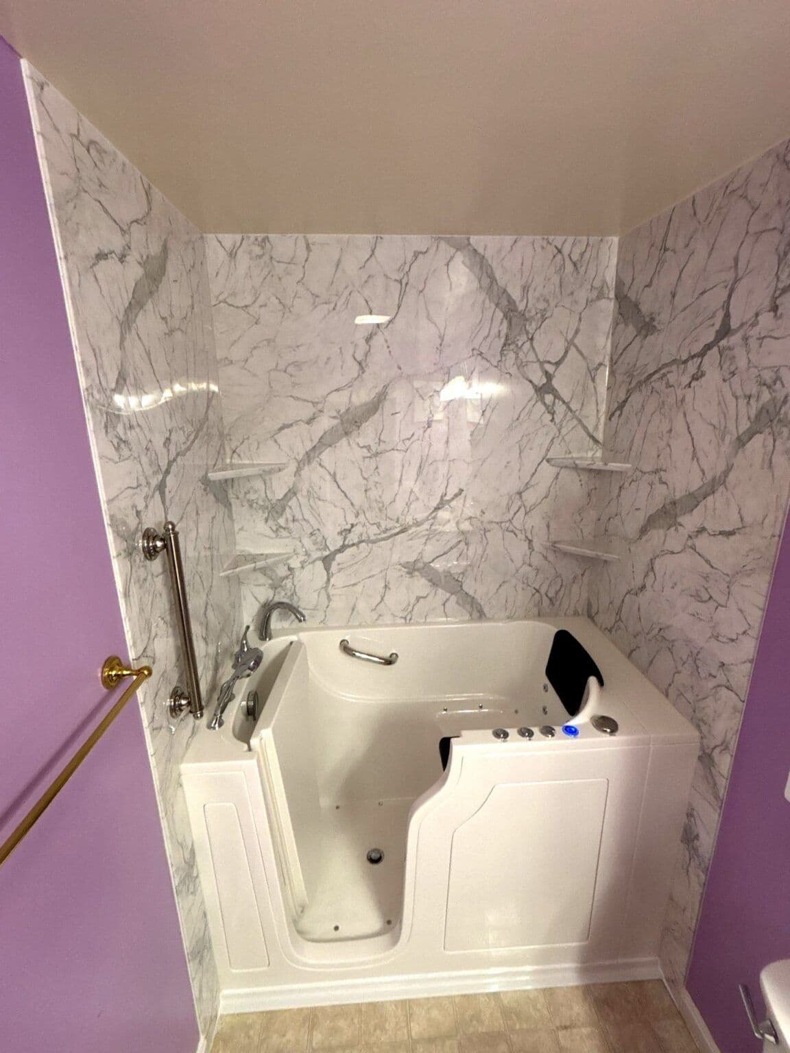 A purple bathroom with a newly-installed walk-in accessible bathtub with grab bar, marbled walls, and corner shelves.